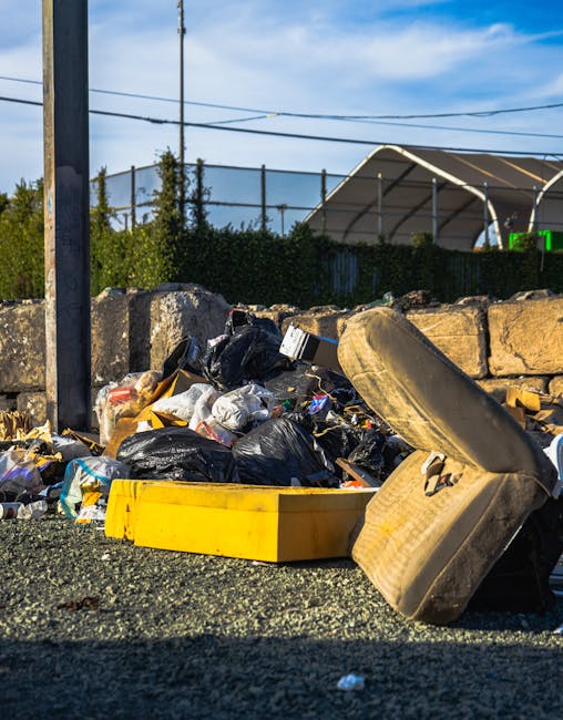 A white garbage collection truck with visible rust and wear on its rear loading compartment is stationed on a narrow urban street lined with older, multi-storey buildings with a mixture of stone and b